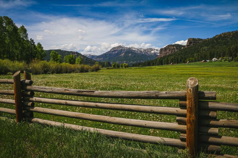 A scenic view of a wooden fence in a lush green field with mountains and a clear blue sky in the background.