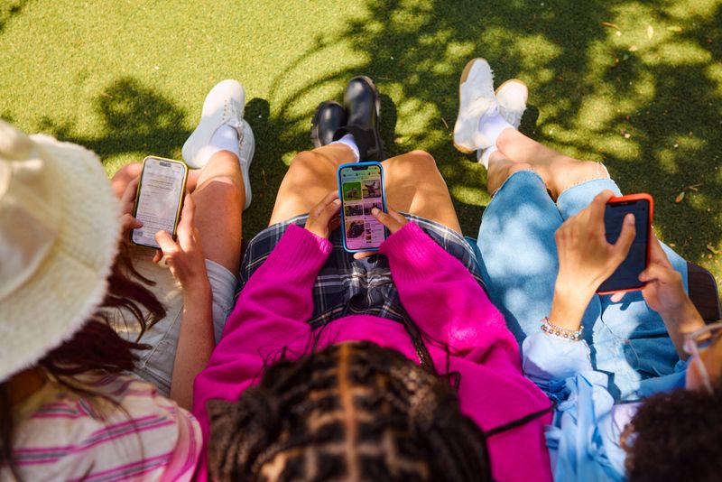 Three friends are using their smartphones while sitting on a bench in a park