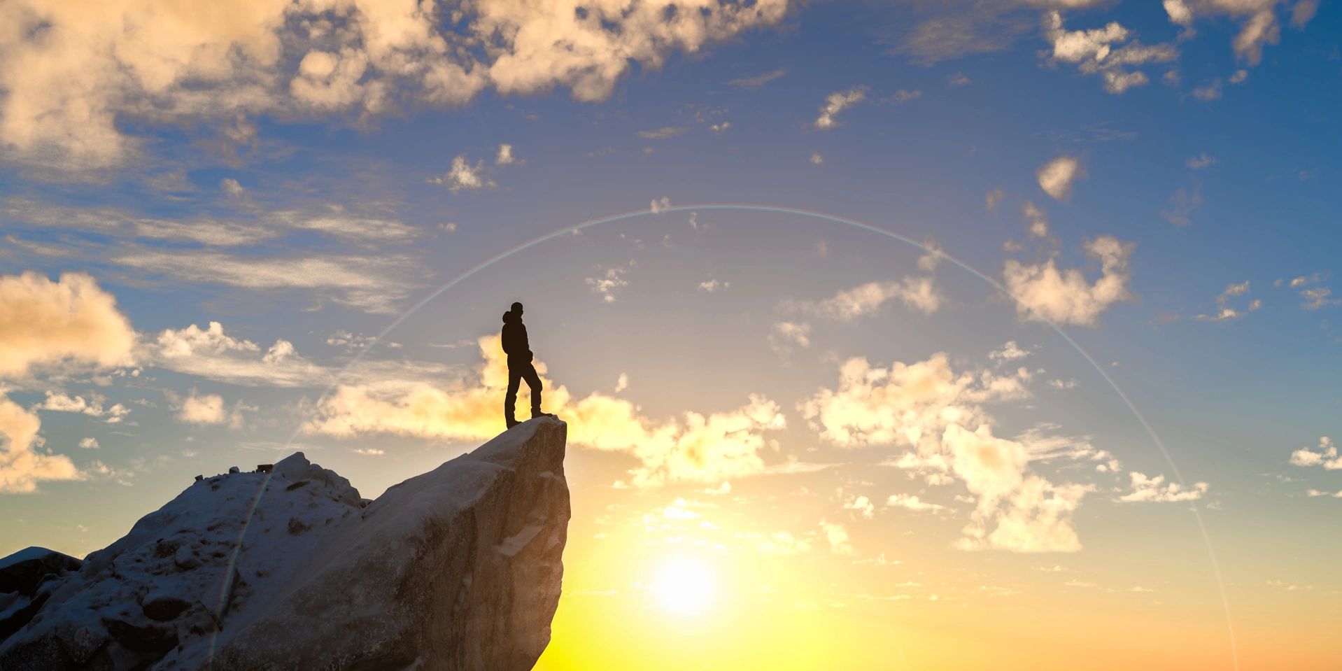 Person stands on a rocky peak at sunset, silhouetted against the colorful sky.