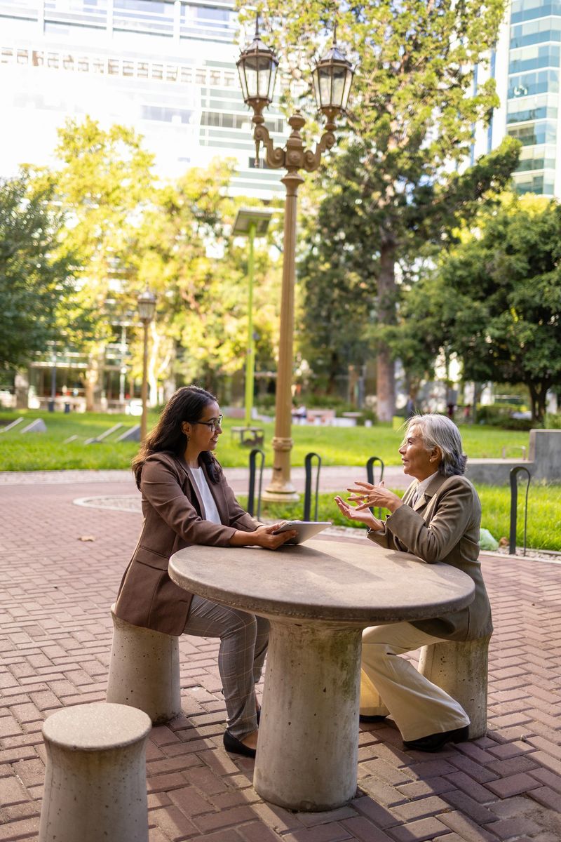 Two businesswomen engage in a conversation, seated at a round outdoor table in a city park.