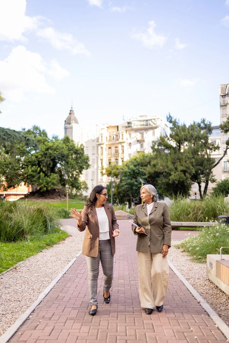 Two professional women walking together in a park while discussing business topics. The background shows buildings and nature, emphasizing the contrast between urban environments and green open spaces.