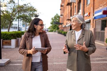 Two women in coats chatting happily outdoors on a city sidewalk.