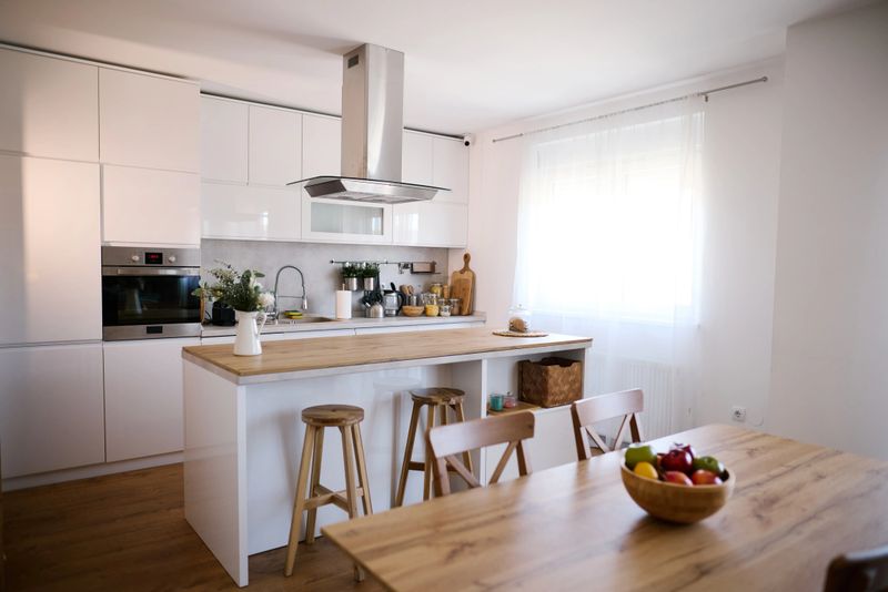 Modern white kitchen with island and wooden table featuring fresh fruit, showcasing a clean and organized space perfect for cooking and dining