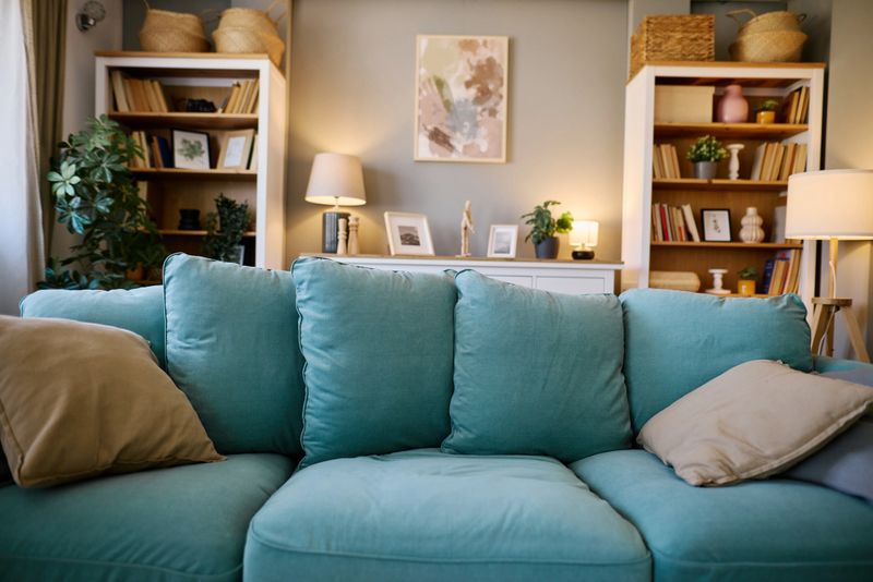 Comfortable turquoise sofa with cushions invites you to relax in a cozy living room, illuminated by soft lamps and decorated with bookshelves and artwork