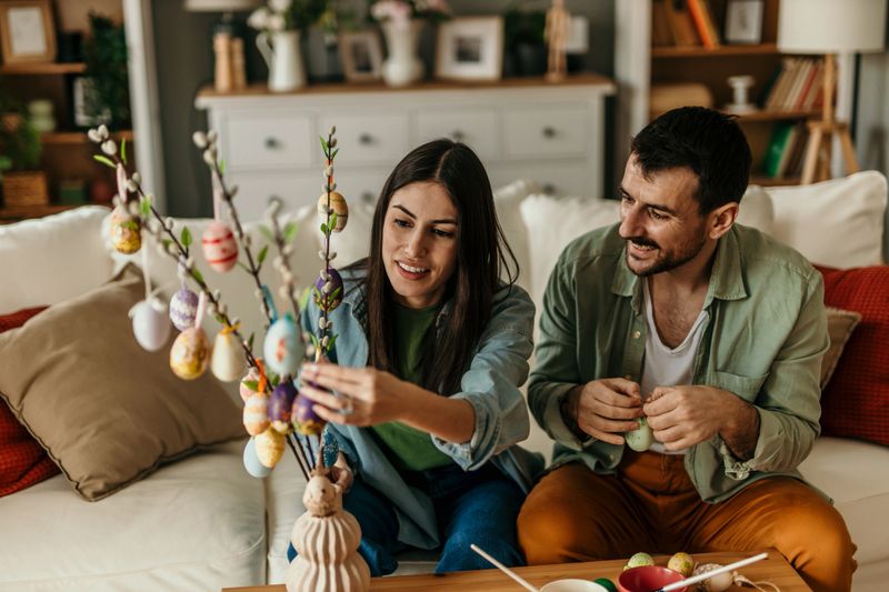 Young couple smiling and decorating colorful Easter eggs and branches together in their cozy living room, enjoying a festive holiday activity