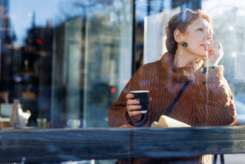 A 48-year-old Caucasian woman with red hair sits alone at a cafe, visible through the window with reflections. She holds a coffee cup in one hand while resting her chin on the other in a thoughtful pose. Wearing a brown sweater, she gazes outside with a contemplative expression. The scene is captured from outside the cafe, showing the window reflection that creates a layered visual effect.
