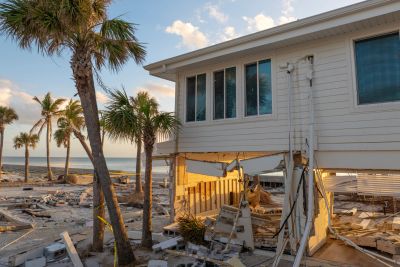 Beach house damaged by storm with debris scattered around.