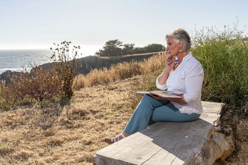 Middle age woman writing in journal outdoors by ocean. Professional model.