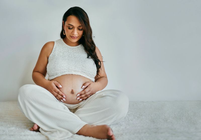 A serene pregnant woman, of diverse ethnicity, sits cross-legged on a textured white surface, wearing soft white attire, gently cradling her belly against a neutral background.