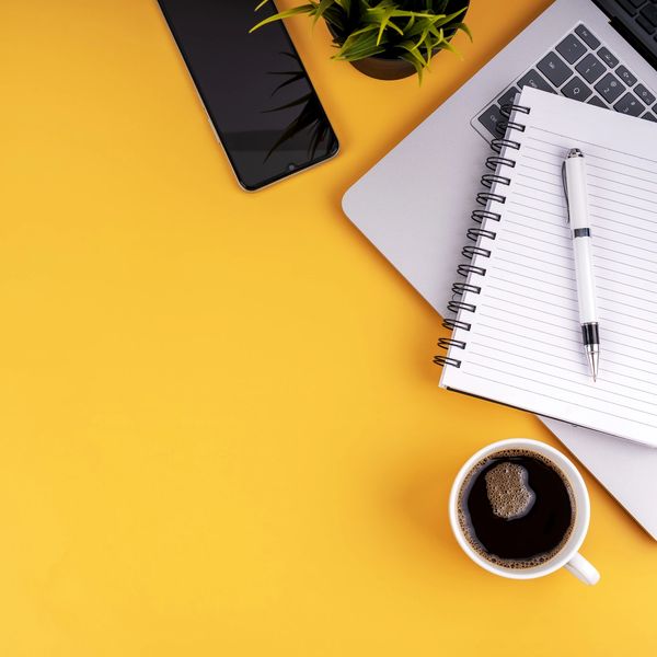 Workspace setup with laptop, notebook, pen, smartphone, plant, and coffee on yellow background.