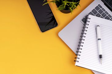 Workspace with phone, laptop, notebook, and pen on a yellow surface.