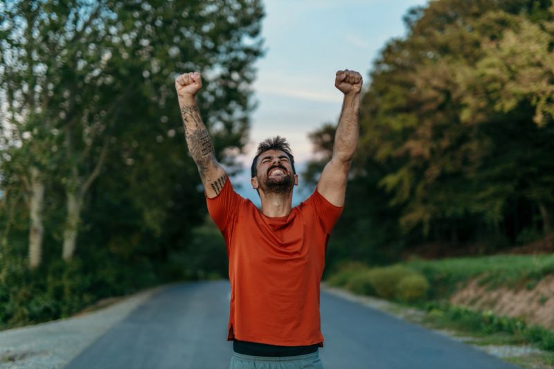 Young sportsman celebrating victory with arms raised, running along a road surrounded by a lush forest, radiating joy and determination