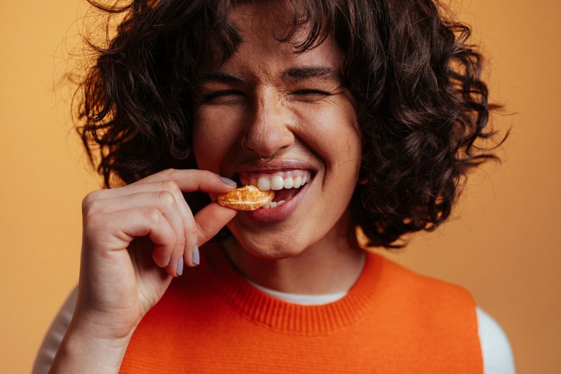 Close-up portrait of a young woman savoring a slice of tangerine with her eyes closed, expressing pleasure and enjoyment