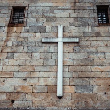 A large white cross on a stone brick wall with two barred windows.