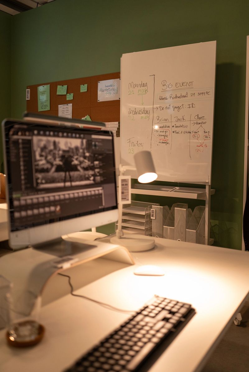 Well-lit, modern office desk features a computer monitor displaying an image, a keyboard, a lamp, and a whiteboard with notes and a schedule