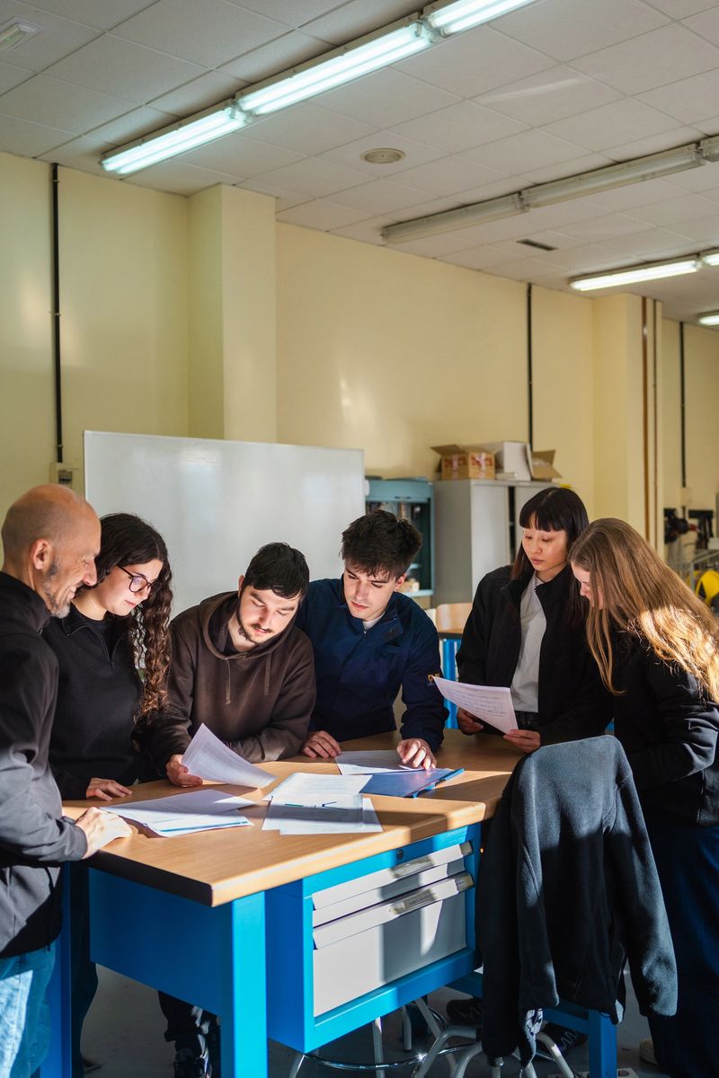 Engineering students with instructor reviewing technical documents, blueprints spread across laboratory workbench during collaborative project planning