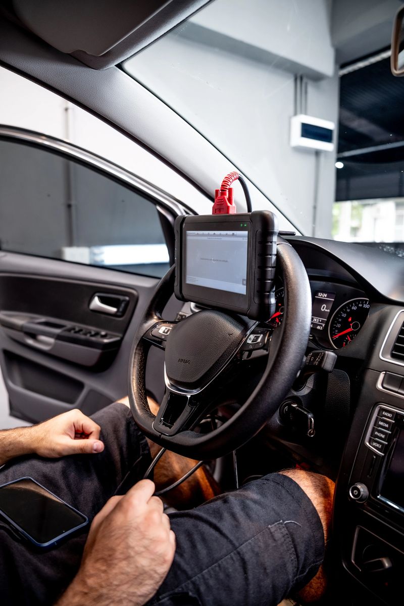 An automotive technician using a diagnostic tool mounted on the steering wheel inside the modern interior of a car, checking technical data and performing system tests on the vehicle.