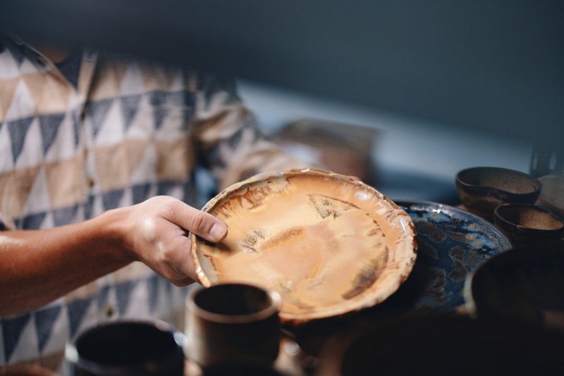 A person holding a rustic, handcrafted ceramic plate above a collection of pottery pieces, showcasing artistic craftsmanship and ceramic art. Ideal for themes of artisanship, pottery, and handmade goods.