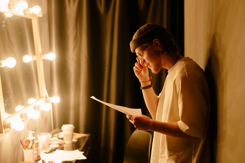 Individual standing backstage holding script next to illuminated mirror reflecting on upcoming performance considering last-minute preparations rehearsing quietly for next scene