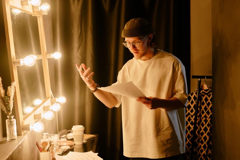 Person standing in front of vanity mirror with lights, holding papers and gesturing. Dramatic shadows and warm lighting enhance performing mood in dressing room