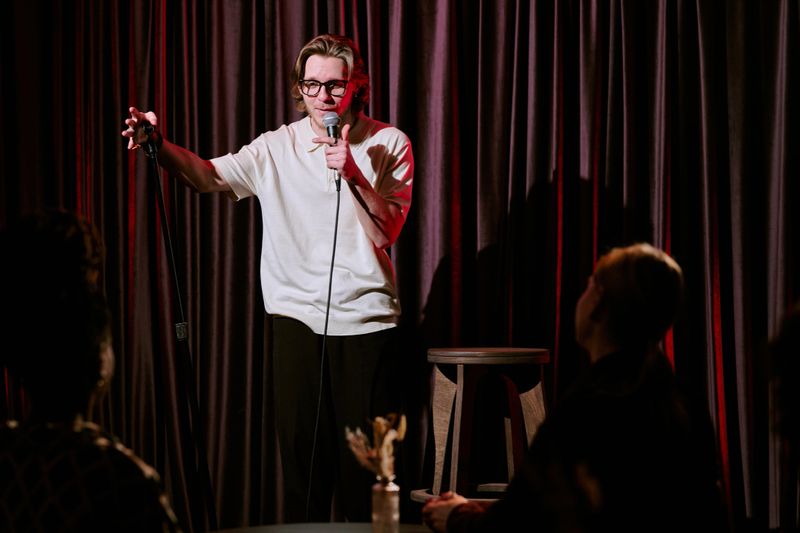 Comedian engaging audience with humor and wit during stand-up performance on stage. Microphone held confidently, small crowd gathered for evening entertainment