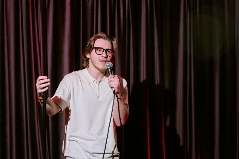 Man engaging audience with humorous performance on stage while holding microphone and curtain in the background
