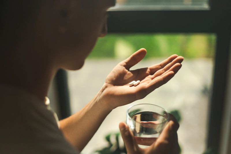 An individual holding pills and a glass of water, preparing medication intake in a bright, reflective environment near a window.