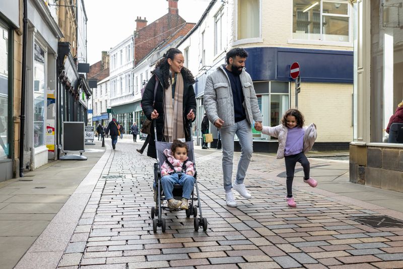 A wide shot of a happy family enjoying a stroll through a pedestrian shopping street. The mother pushes a stroller with a young child, while the father holds hands with their older daughter. The scene captures a warm and joyful family moment. Located in Hexham, Northumberland.Videos of this scenario are available