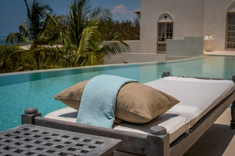 A cushioned lounge chair sits beside a serene swimming pool in a tropical setting, with palm trees and a luxury villa in the background. Soft blue and beige towels are placed neatly, evoking a tranquil atmosphere perfect for vacation, spa, or luxury lifestyle concepts. No people are present in the scene.
