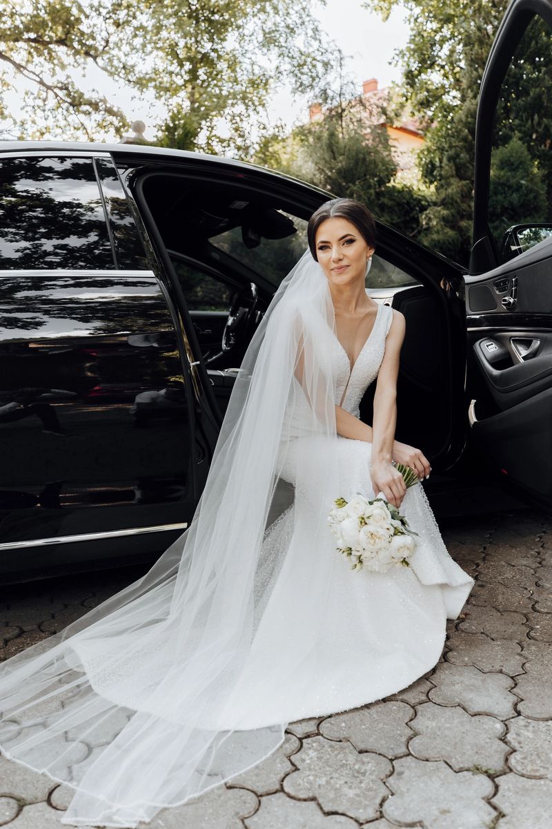 A bride is sitting in the back of a black car with a white veil. She is holding a bouquet of flowers