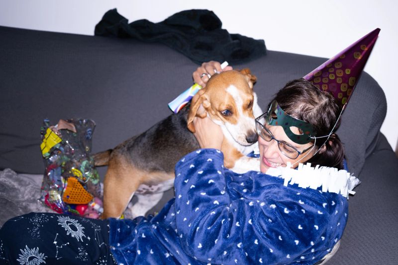 Mature woman wearing pajamas, a party hat, and a mask enjoys a playful moment with her beagle on a sofa, surrounded by party favors, celebrating at home
