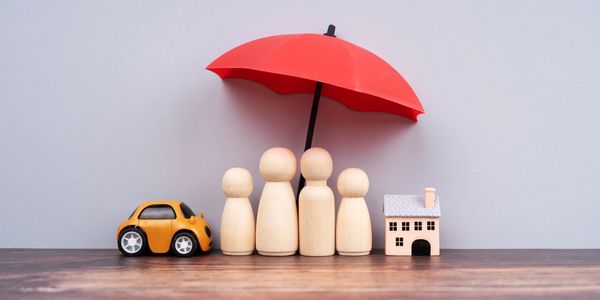 Wooden family figures protected by a red umbrella, with a toy car and house beside them.