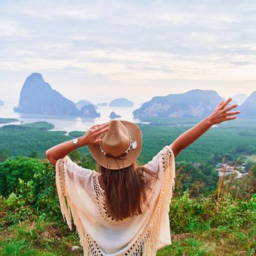 Woman with hat enjoying scenic mountain and water view with arms raised.