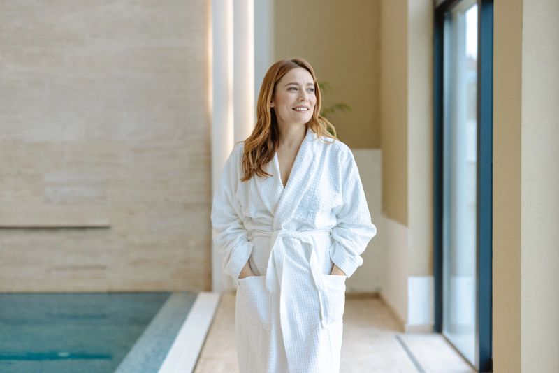 A serene woman wearing a white robe, enjoying the view near an indoor spa pool. Perfect for concepts of relaxation, wellness, self-care, and rejuvenation in a calm and luxurious setting.