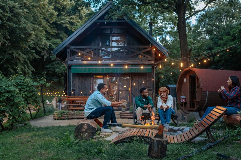 Group of friends relaxing and chatting around bonfire in front of their cabin in the woods, enjoying their vacation