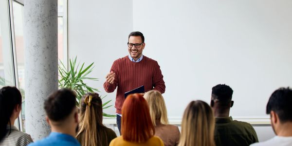 Smiling man giving a presentation to an attentive audience in a bright room.