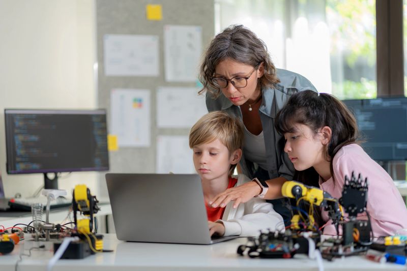 A skilled female teacher sits with students, explaining how to design and program a robotic arm in a hands-on STEM lesson. This engaging classroom environment fosters creativity, problem-solving, and expertise in robotics, artificial intelligence, and coding.