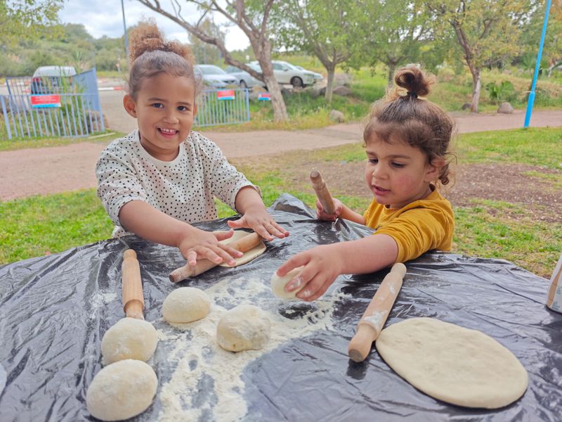 A young girl and a boy are playing together at an outdoor table, rolling dough with wooden rolling pins. They are surrounded by small dough balls and flour on a black table cover, appearing focused and happy while engaging in a fun and educational cooking activity in a park setting.