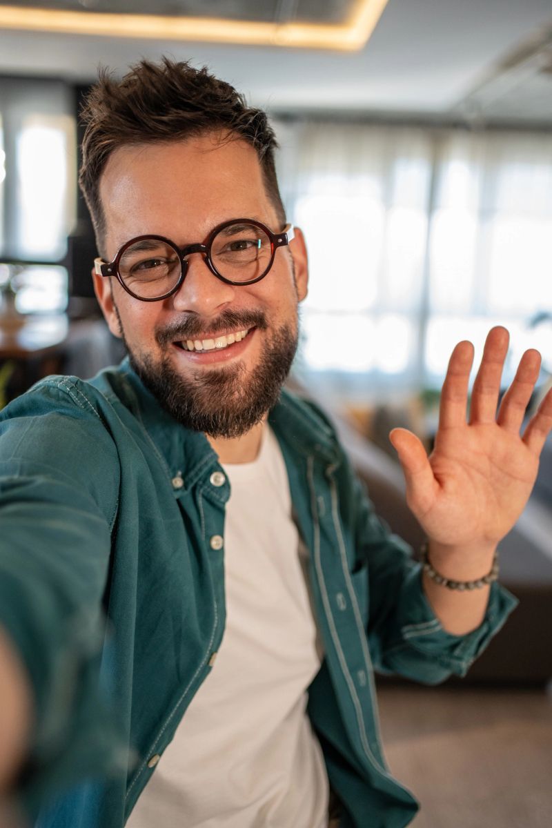 Young, bearded man with glasses smiling and waving while making a video call from home, capturing a joyful moment with a selfie in a casual and inviting living room setting
