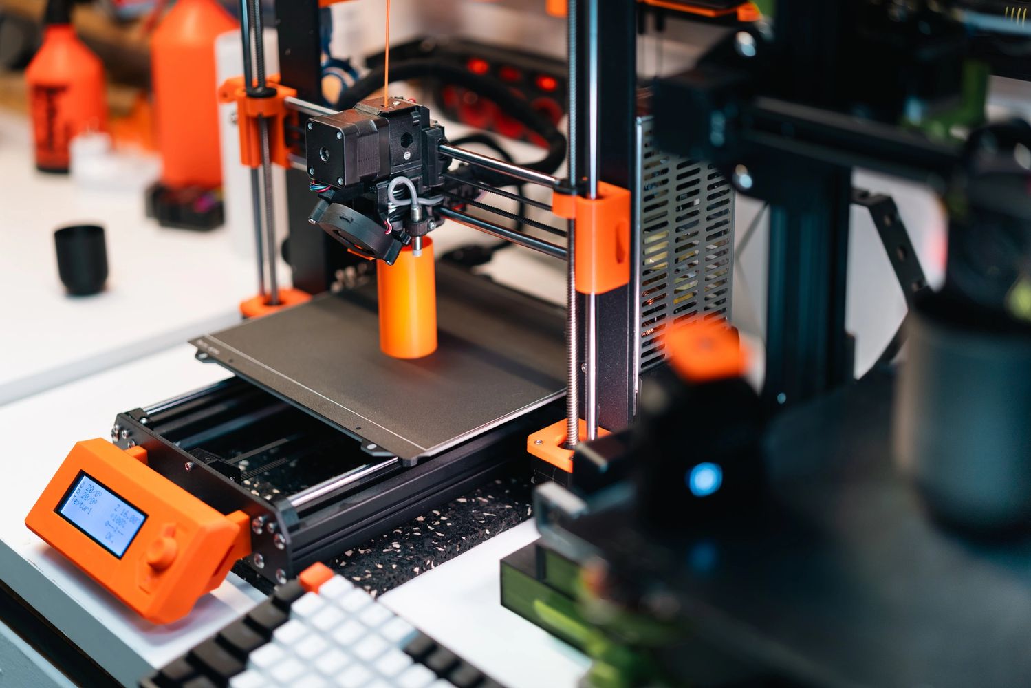 3D printer creating an orange cylindrical object on a workbench.