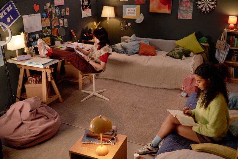 Two friends are studying in a cozy, well-decorated bedroom filled with personal items. One is seated at a desk while the other is reading on a bed, creating a warm and focused atmosphere