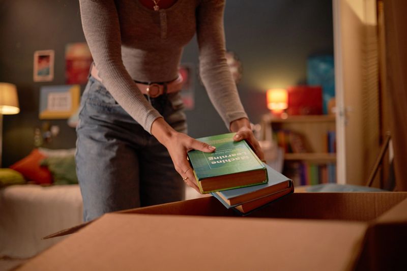 Person placing books into cardboard box in cozy room with shelves, decor, and warm lighting creating inviting atmosphere for organizing or relocating items
