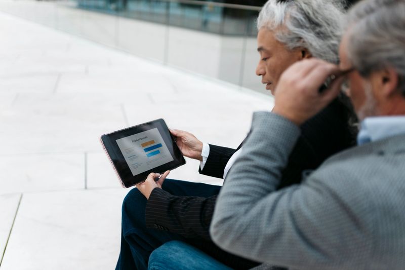 Two business people are reviewing product growth data on a digital tablet, discussing strategy and performance in a modern outdoor setting