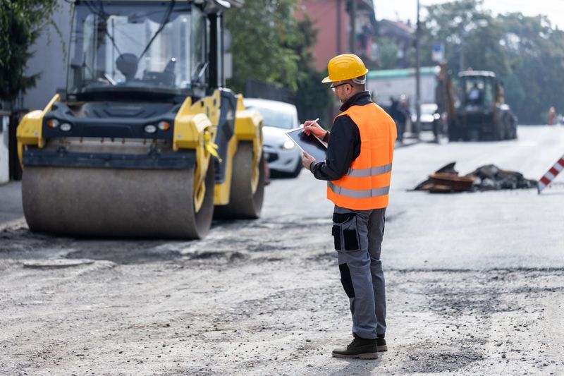 A construction worker wearing a helmet and reflective vest inspects a road construction site. He checks details on a clipboard while large machinery operates nearby, indicating ongoing repairs.