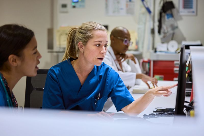 Nurse pointing at computer screen while discussing patient care with colleague. Medical staff working together in busy hospital setting. Healthcare professionals focused on providing best treatment.