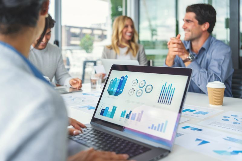 Close up of a digital tablet with data and finance charts and graphs. Group of happy business people in a meeting in then board room in the background. There are laptops and other technology on the table along with documents showing financial data