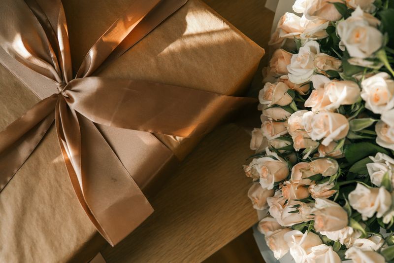 Close-up of a wrapped gift with a satin ribbon next to a bouquet of delicate pale pink roses in soft natural light.