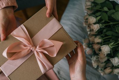 Two hands exchanging a gift box with a pink ribbon beside a bouquet of white roses.