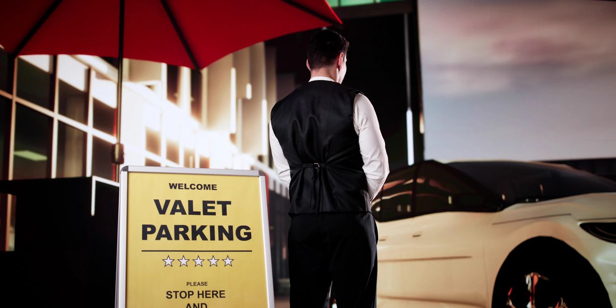 A valet attendant stands near a yellow valet parking sign and a white car under a red umbrella.
