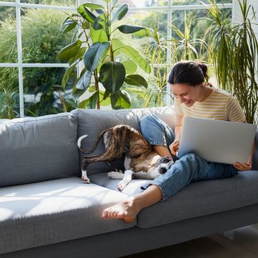 A woman laughing with her dog on a couch by a sunny window.